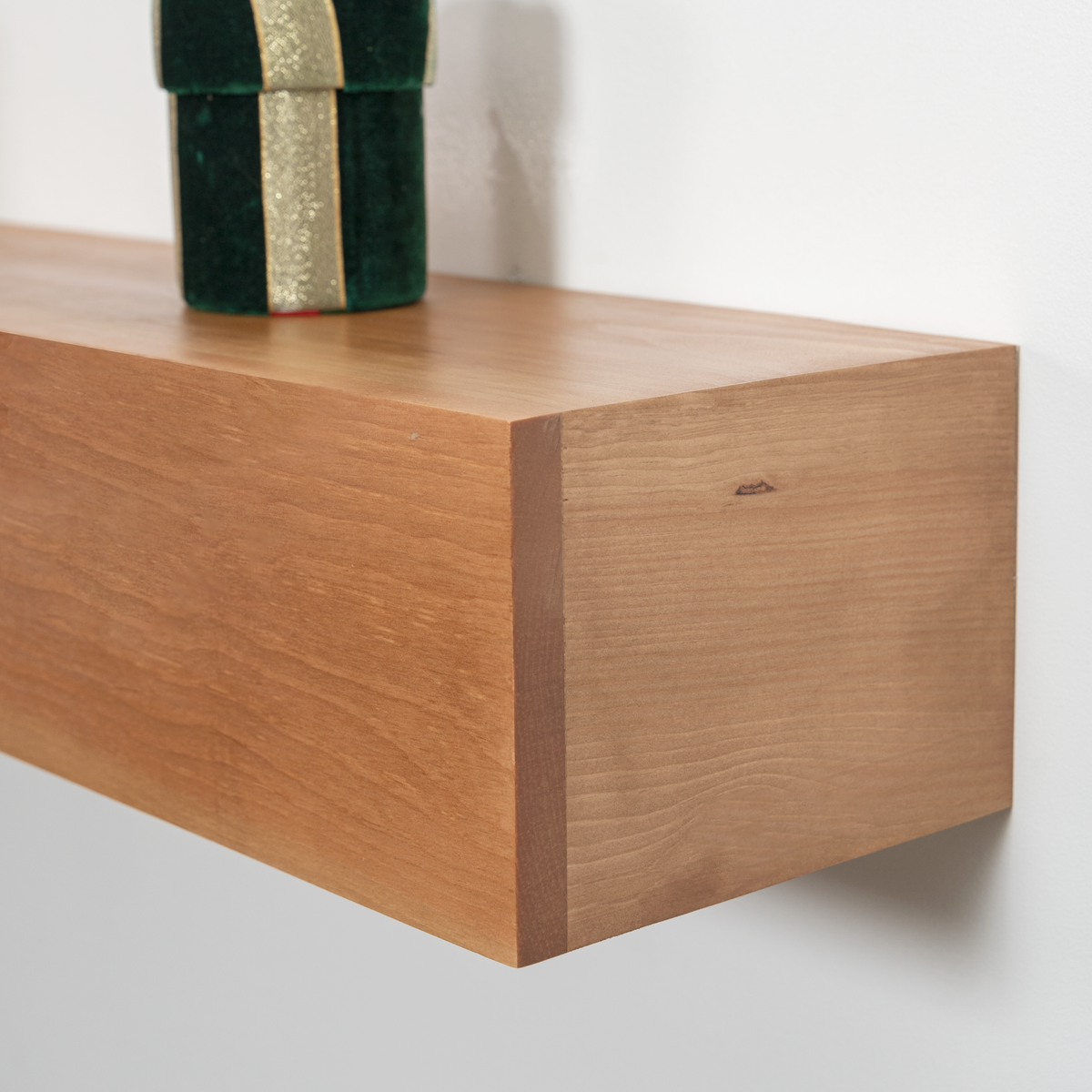 A close-up of a light brown wooden floating fireplace mantel with a visible grain texture, mounted on a white wall. A dark green gift box with a gold ribbon is partially visible on top of the shelf.
