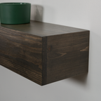 Dark brown wooden floating fireplace mantel with visible wood grain, mounted on a light gray wall with a green planter on top.
