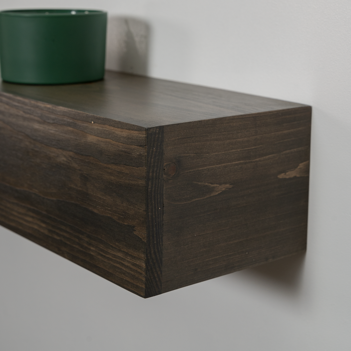 Dark brown wooden floating fireplace mantel with visible wood grain, mounted on a light gray wall with a green planter on top.