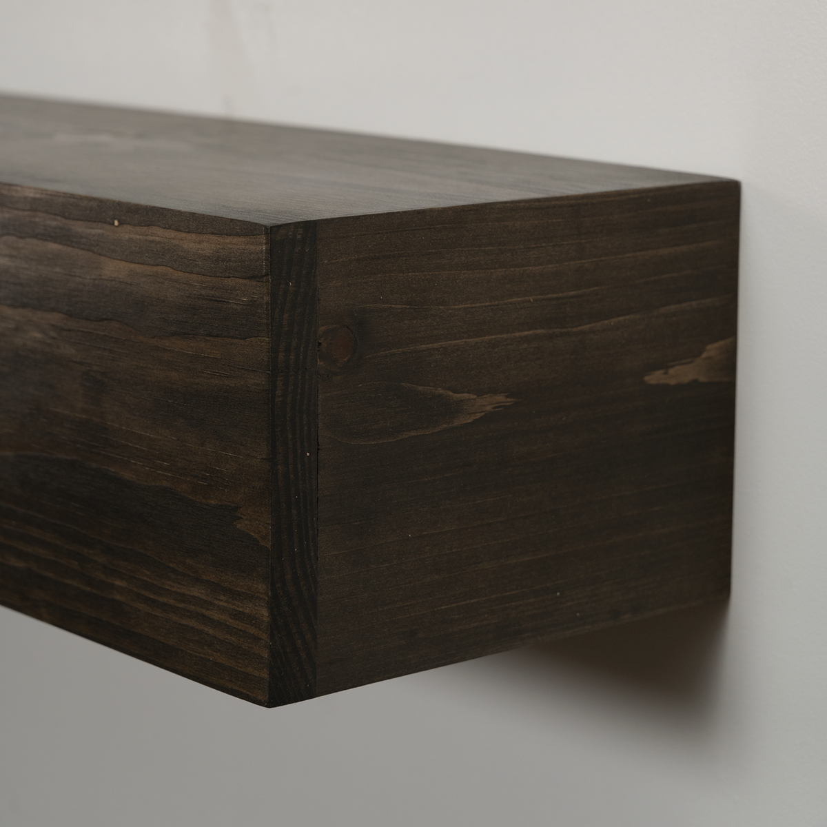 Dark brown wooden floating fireplace mantel with visible wood grain and a wall-mounted corner, shown against a plain light gray wall.