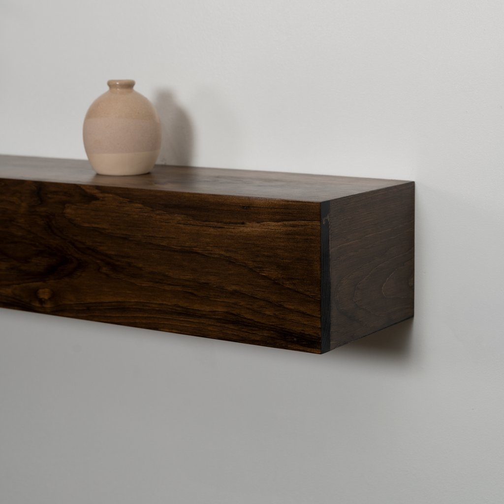 A dark brown floating wooden fireplace mantel mounted on a light gray wall, with a small beige ceramic vase placed on top.