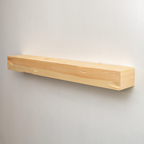 A minimalist, long rectangular wooden floating fireplace mantel in a light natural pine finish, mounted on a plain light gray wall.