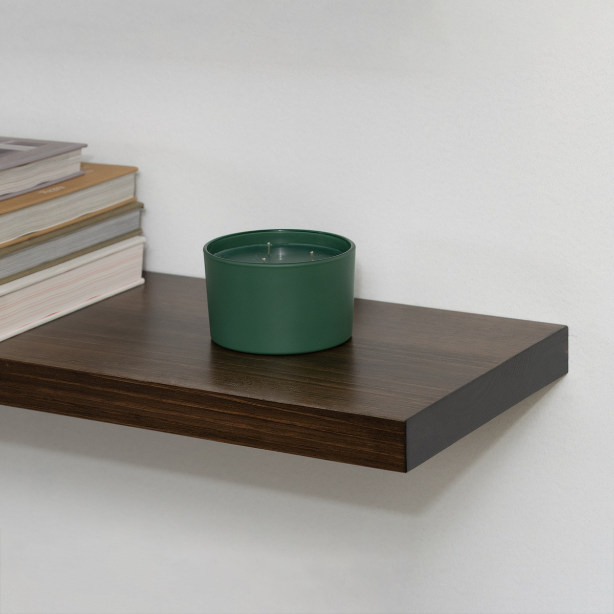 Green pot on a wooden floating shelf with books stacked beside it against a white wall.