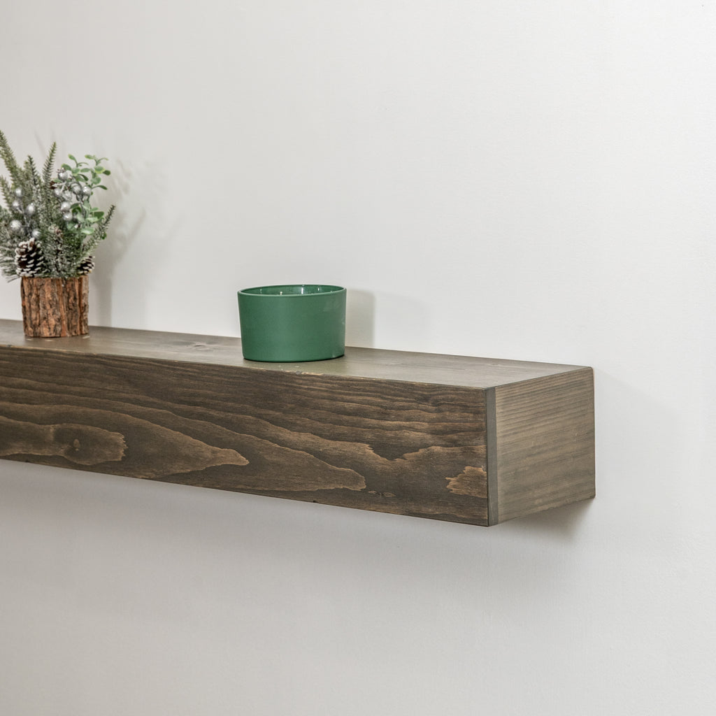 A rustic dark brown wooden floating fireplace mantel mounted on a white wall, styled with a small green ceramic cup and a potted pine branch.