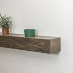 A rustic dark brown wooden floating fireplace mantel mounted on a white wall, styled with a small green ceramic cup and a potted pine branch.
