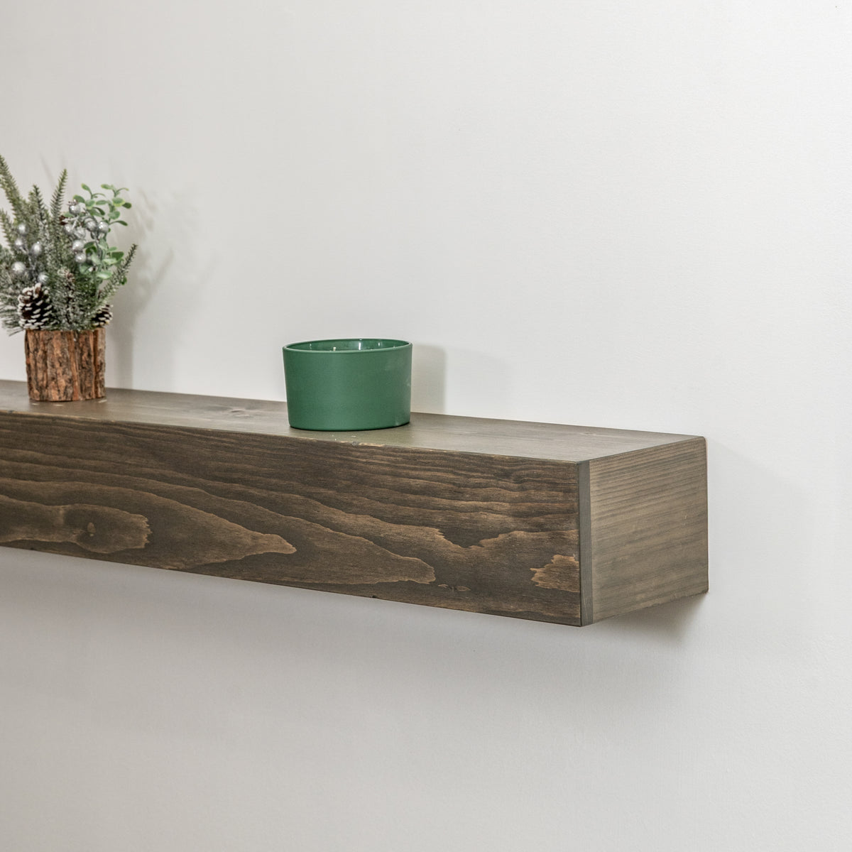 A rustic dark brown wooden floating fireplace mantel mounted on a white wall, styled with a small green ceramic cup and a potted pine branch.
