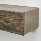 A close-up of a floating wooden fireplace mantel with a dark brown wood grain finish, mounted against a plain white wall.