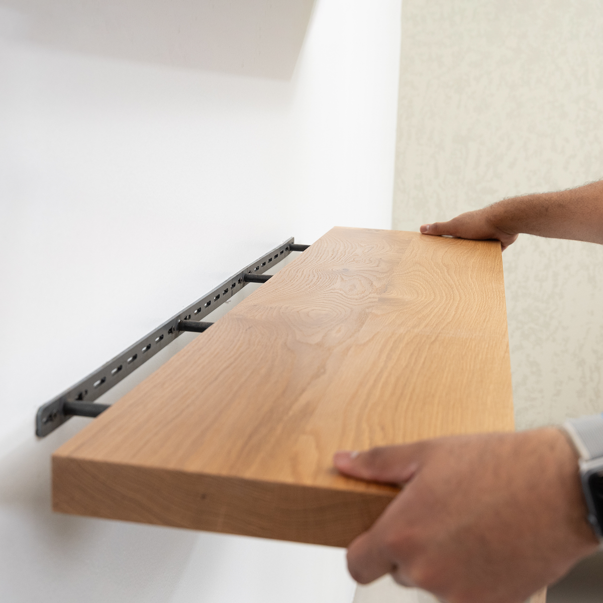 Wooden floating shelf being placed on a metal bracket by a person against a white wall.