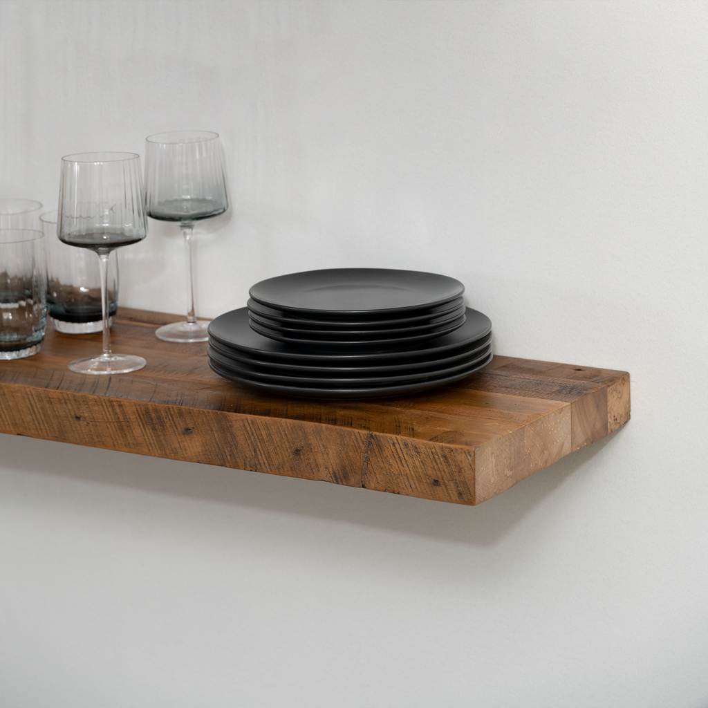 Stack of black plates on a wooden floating  shelf with wine glasses against a white wall