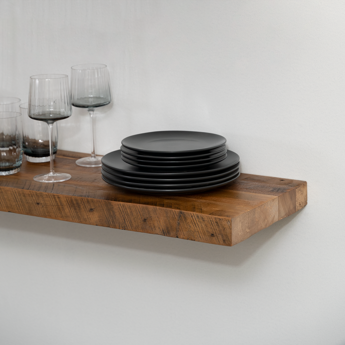 Stack of black plates on a wooden floating  shelf with wine glasses against a white wall