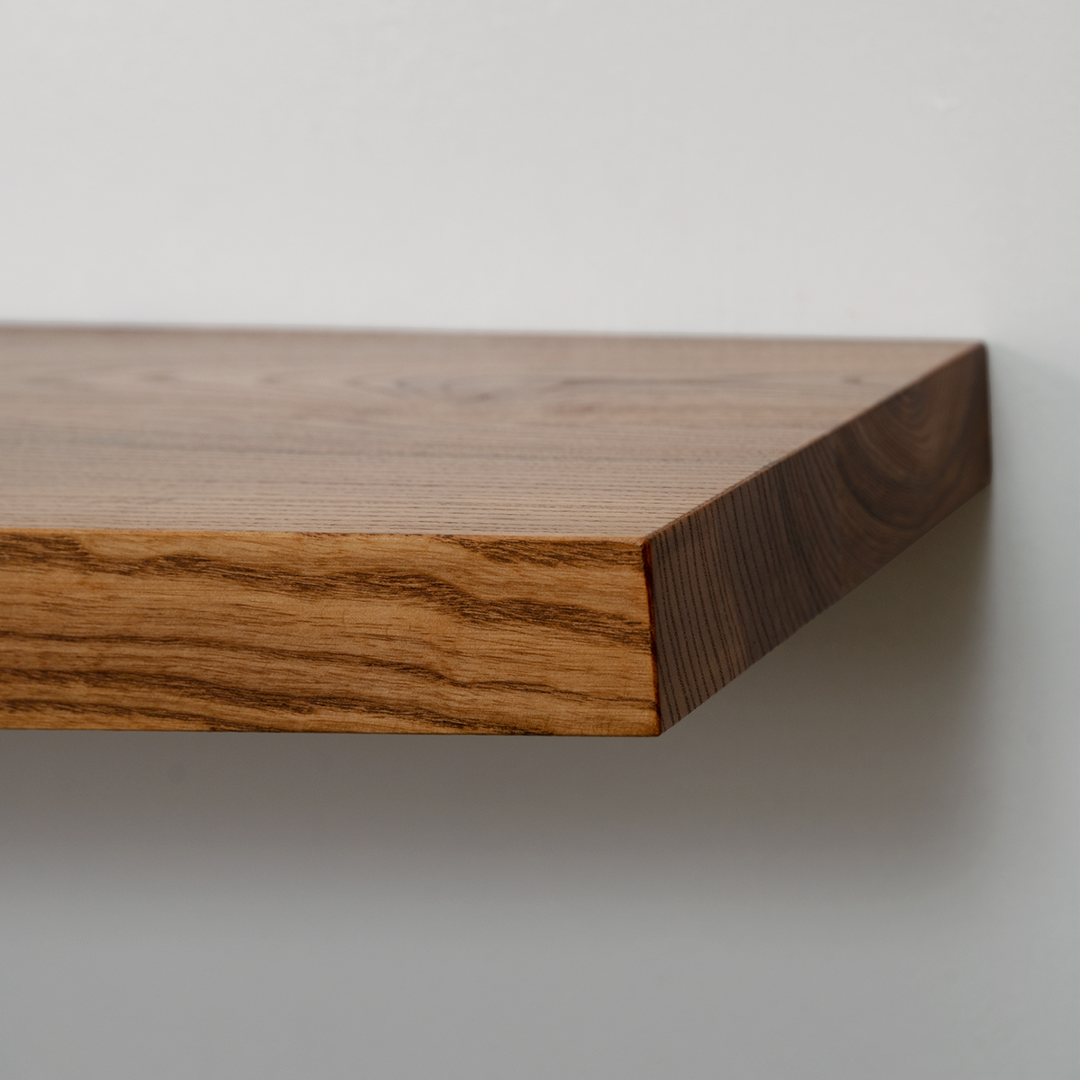A close-up of a floating wooden shelf with a rich medium-brown wood grain finish, mounted against a plain light gray wall.