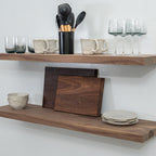 Wooden floating shelves with kitchen items including mugs, glasses, and cutting boards on a white wall.