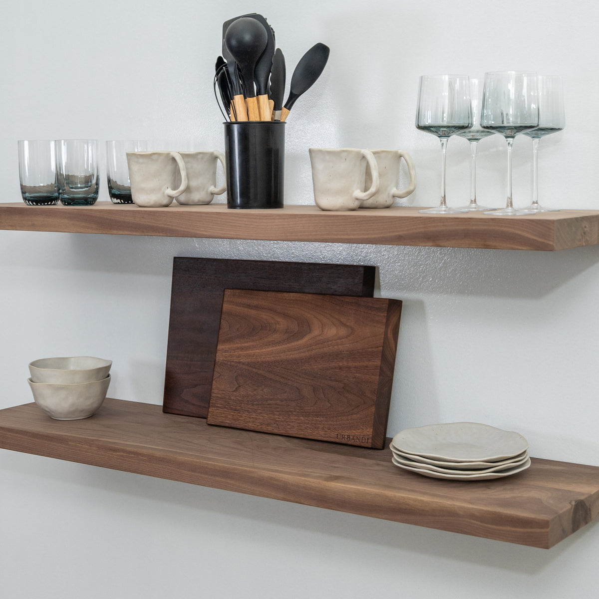 Wooden floating shelves with kitchen items including mugs, glasses, and cutting boards on a white wall.