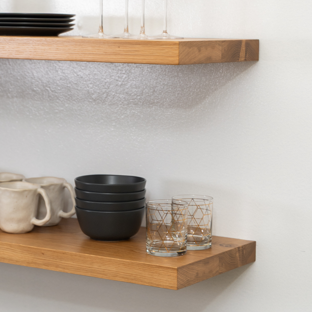 Wooden white oak floating shelves with ceramic mugs, black bowls, and glass tumblers against a white wall.