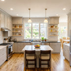 A bright, modern kitchen with white shaker-style cabinets, a central island with two dark wood bar stools, a double sink, and a large window looking out onto a backyard.